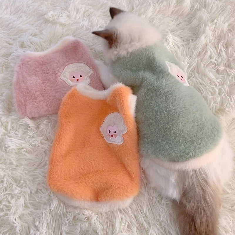 Three colorful pet vests with cartoon faces on a fluffy white surface.