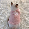 Small dog wearing a pink fluffy sweater with a cartoon face on a white fluffy surface