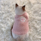 Small dog wearing a pink fluffy sweater with a cartoon face on a white fluffy surface